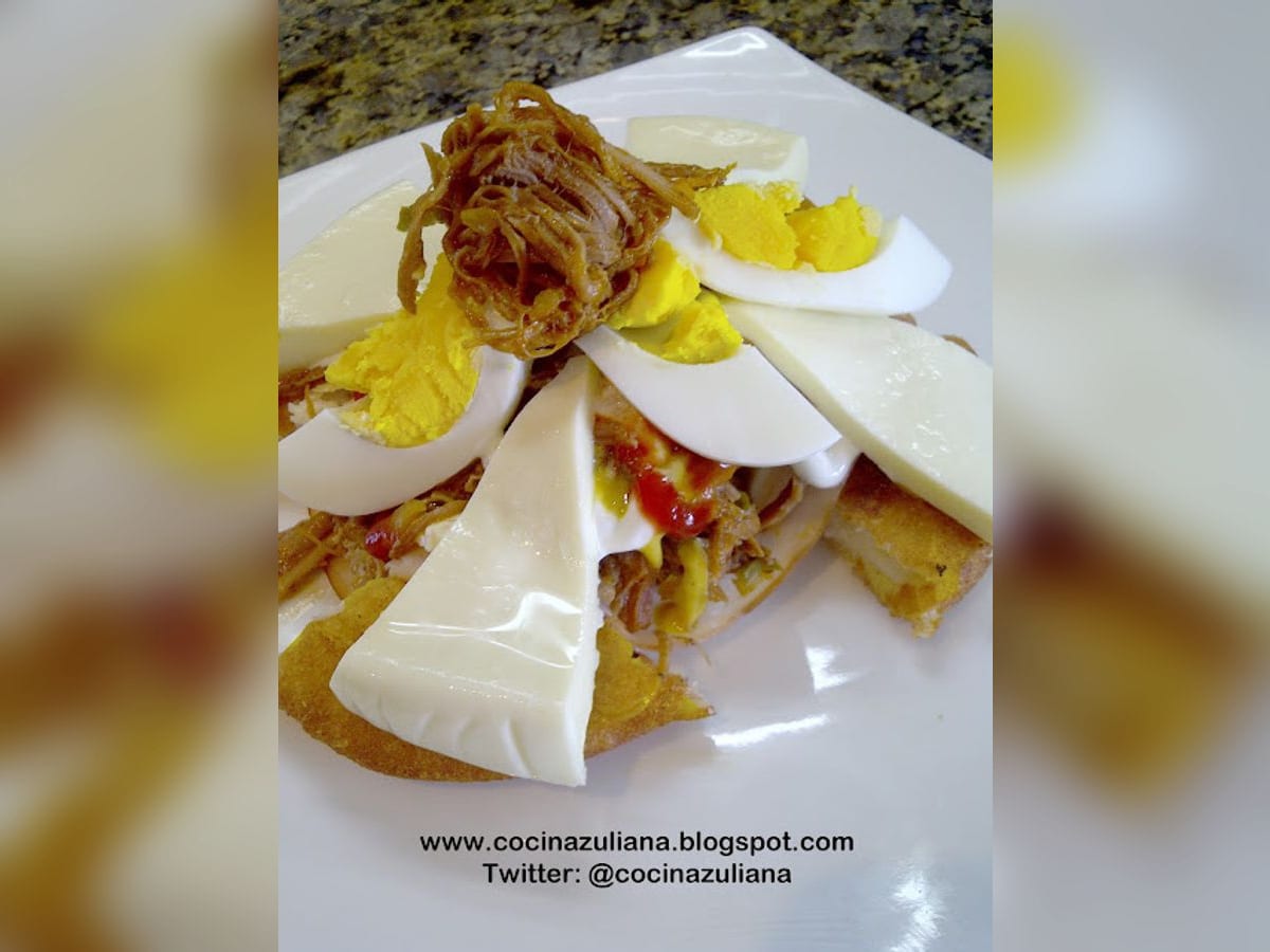 Arepa cabimera frita cortada en porciones, cubierta con carne mechada, jamón, queso, huevo y salsas